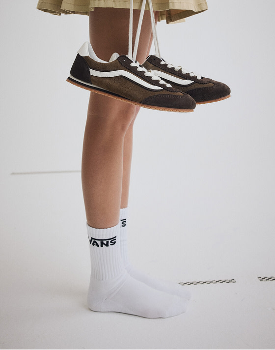 Mid thigh down shot of a woman wearing a pair of White Classic Crew Socks while holding a pair of Demitasse Brown Super Lowpro shoes by the laces.