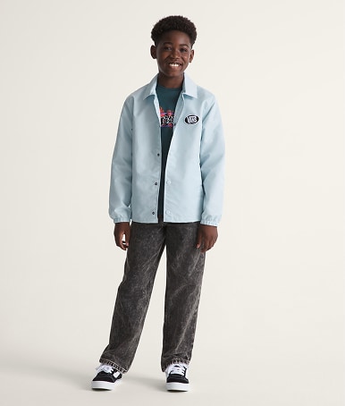 A young boy poses in a photography studio wearing a dawn mist blue Kids Torrey Coaches Jacket with black and white Knu Skool.