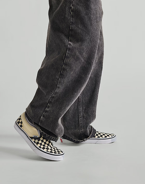 ​​Knee down shot of a kid wearing black jeans and a pair of the black and white Little Kids Classic Slip-On Checkerboard Shoe in a grey photography studio.