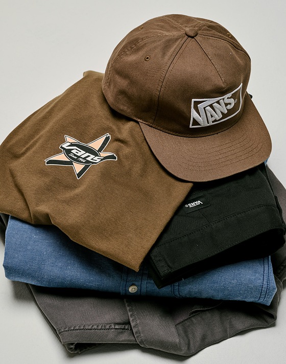 A stack of new Vans pants, denim shirts, t-shirts, and hats on the floor of a photography studio.