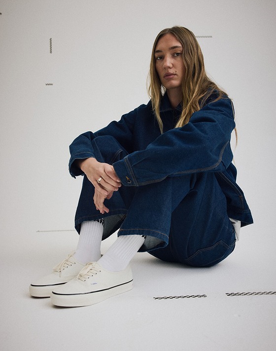 A young woman sits on the floor of a photography studio wearing white Authentic and a dark rise Emily Boxy Work Wear Jacket.