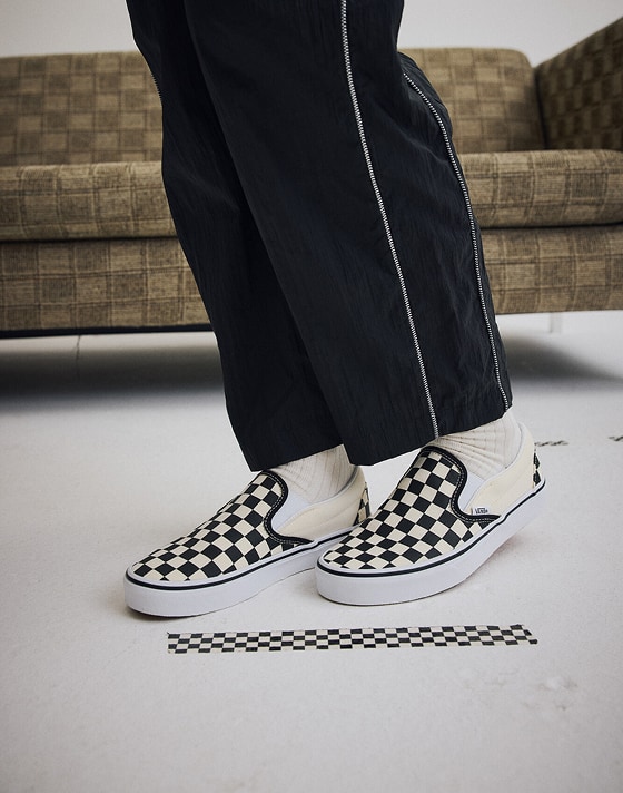 Medium shot knee down of someone standing in a grey photography studio wearing a pair of Black / White Classic Slip-On Checkerboard Shoes.