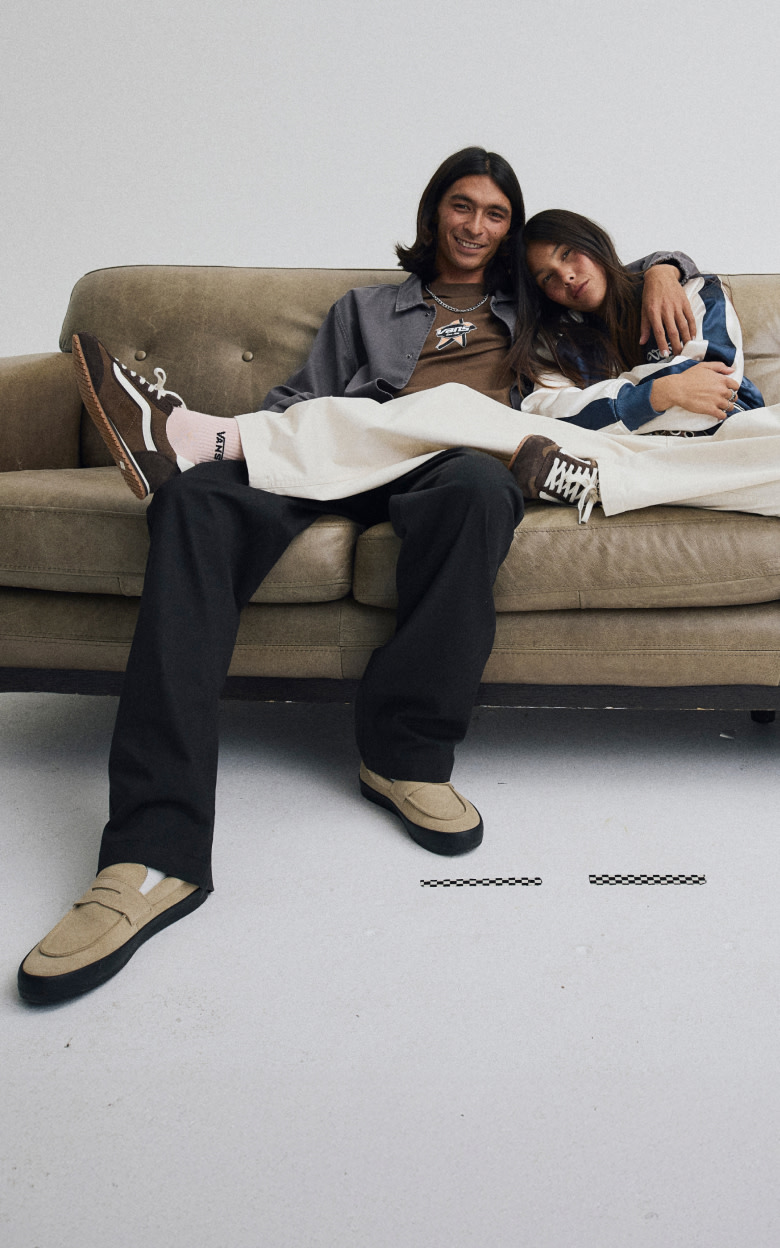 A young man and woman cozy up on a couch together in new Vans clothes, brown Super Lowpro, and tan Skate Loafer shoes.