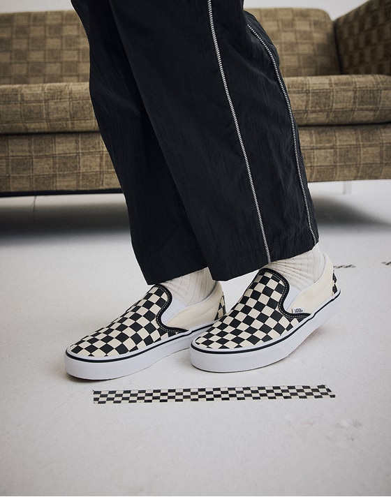 Knee down shot of someone wearing the Nyla Track Pant and a pair of the black and white Classic Slip-On Checkerboard Shoe in a grey studio.