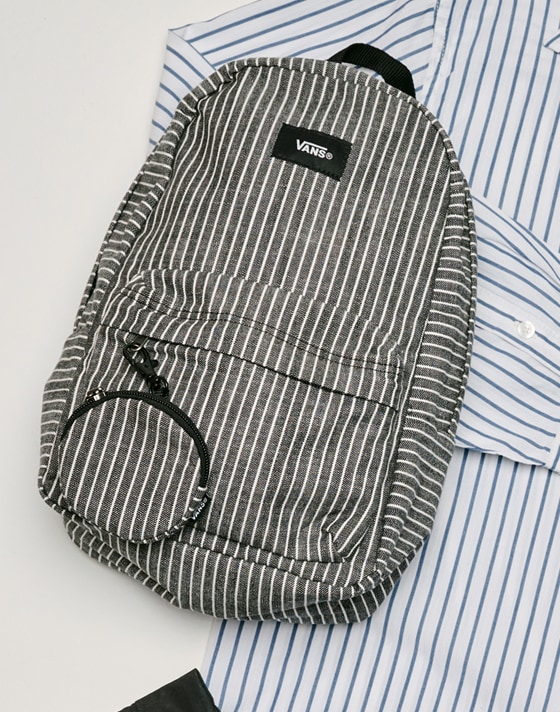 Medium shot of the Old Skool Mini Backpack in white / black laying on top of﻿ a striped shirt on the floor of a grey photography studio.