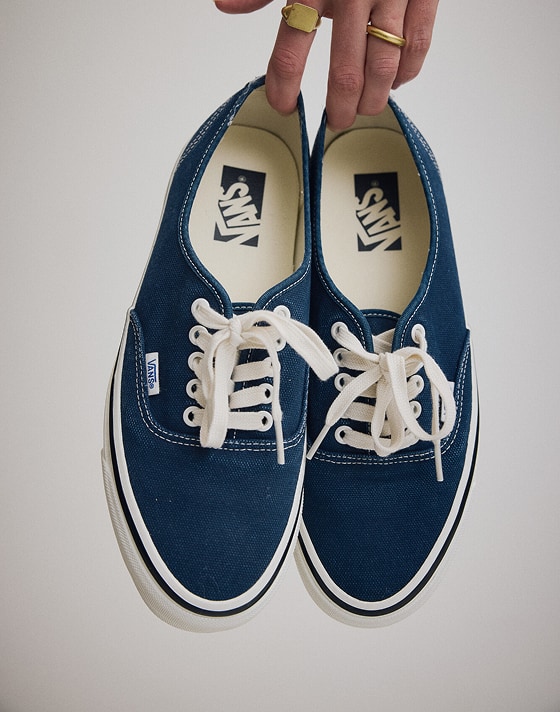 Close up of the Premium Authentic Shoe in Parisian Night Blue dangling from the fingertips of a person in a grey photography studio.