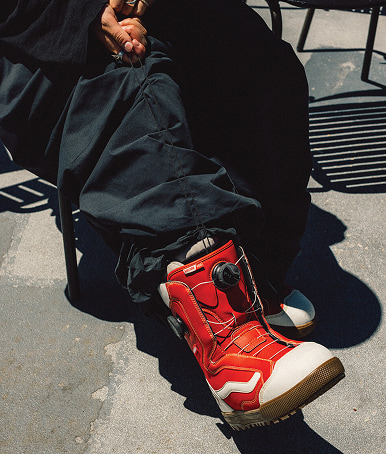 Shot of a person sitting on a chair outside wearing black pants and Women's Encore Pro X Mia Brookes Snowboard Boot in racing red and white.