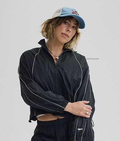 Medium shot of a woman wearing a Black Nyla Tracksuit and a Marked Jockey Trucker Hat in a grey photography studio.