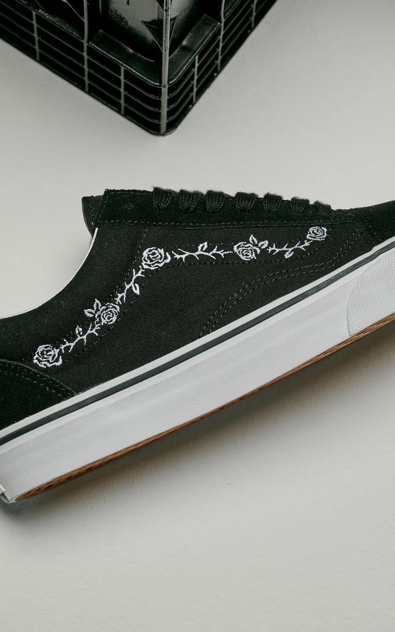 Plan rapproché du Old Skool noir et blanc avec un design de roses et d’épines brodées, posé sur le sol d’un studio photo gris.