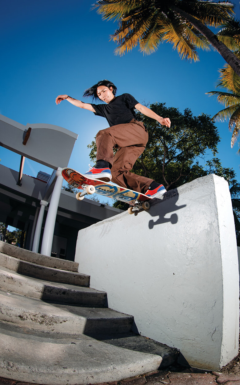 Panoramic shot of Daiki Hoshino outside skateboarding mid jump from a cement ramp over grass onto a driveway wearing the Blue / Red Skate Era Shoe.