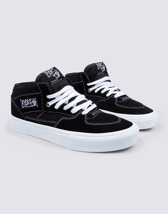 Medium shot of a pair of the Black / White Skate Half Cab Shoes on the floor of a grey photography studio.