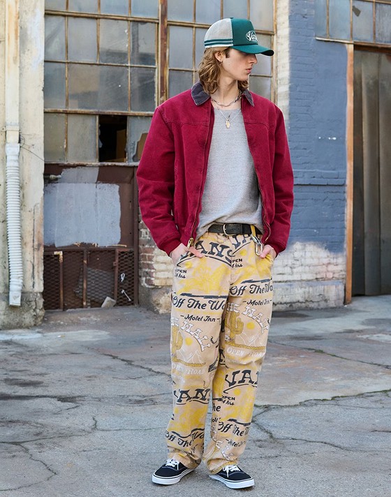 Medium shot of a man outside of a run down building wearing a green Vans hat, red barn jacket, wild west Vans pants, and Black/White Authentic Shoes.
