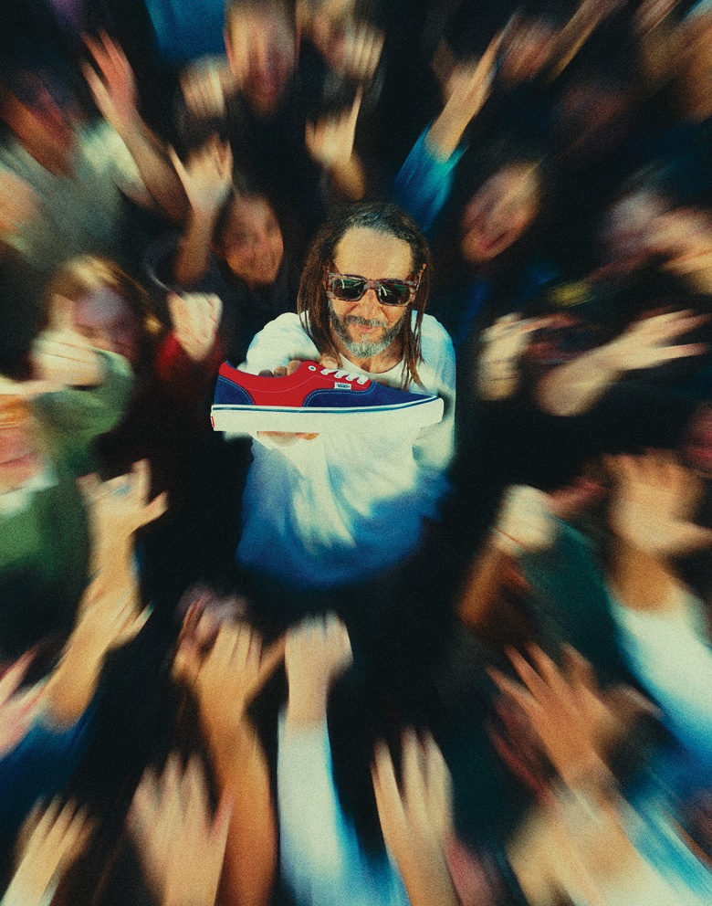 Wide shot of Tony Alva in the middle of a blurred out crowd holding up a Blue / Red Skate Era Shoe that’s in focus.