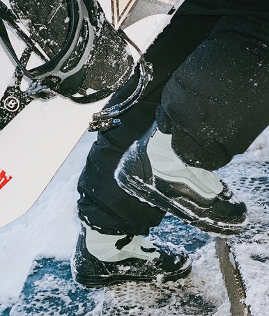 Close-up of someone wearing the olive gray Infuse Snowboard Boot going up snowy steps and holding a snowboard.