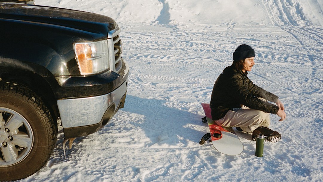 Snowboarder sits on his snowboard in a snowy parking lot in front of his truck.