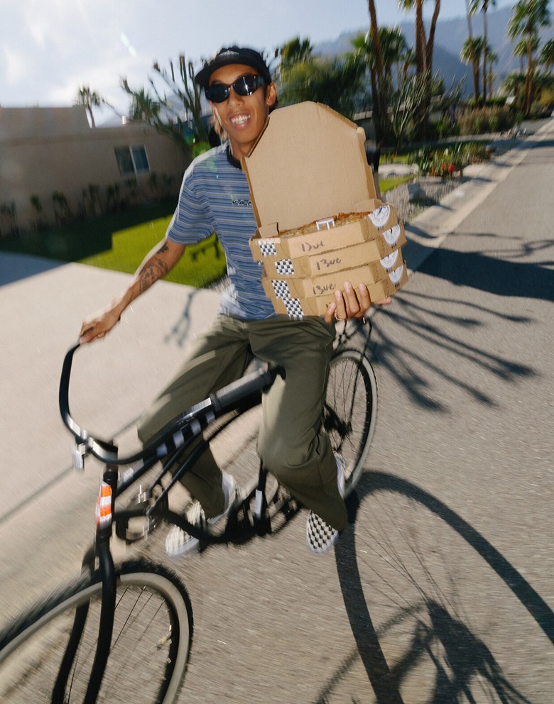 A young man biking outside in the sun wearing Vans clothes holding four pizzas.