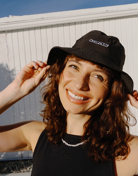 Close-up on a young woman smiling in the sun wearing a faded black Dropped V Bucket Hat.