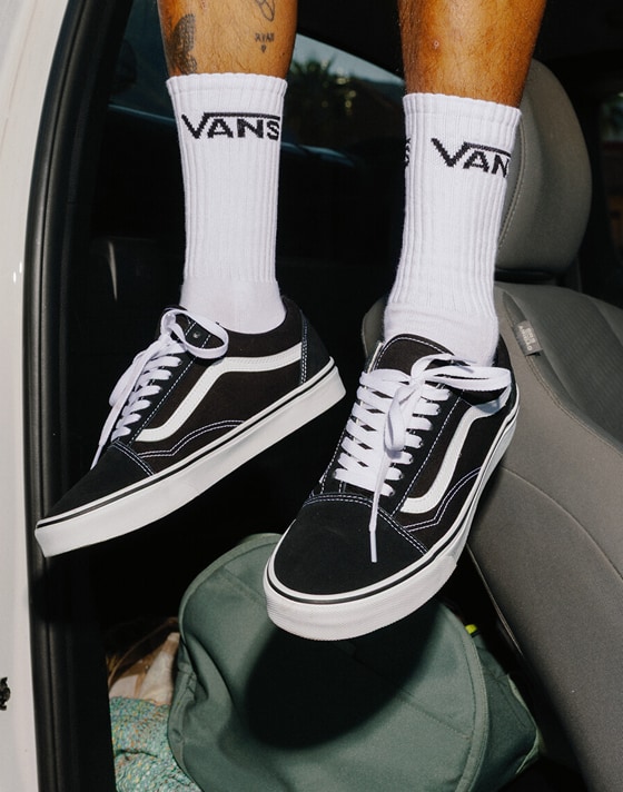 A knee-down shot of someone sitting on the cab of a truck wearing black and white Old Skool shoes and white Vans crew socks.
