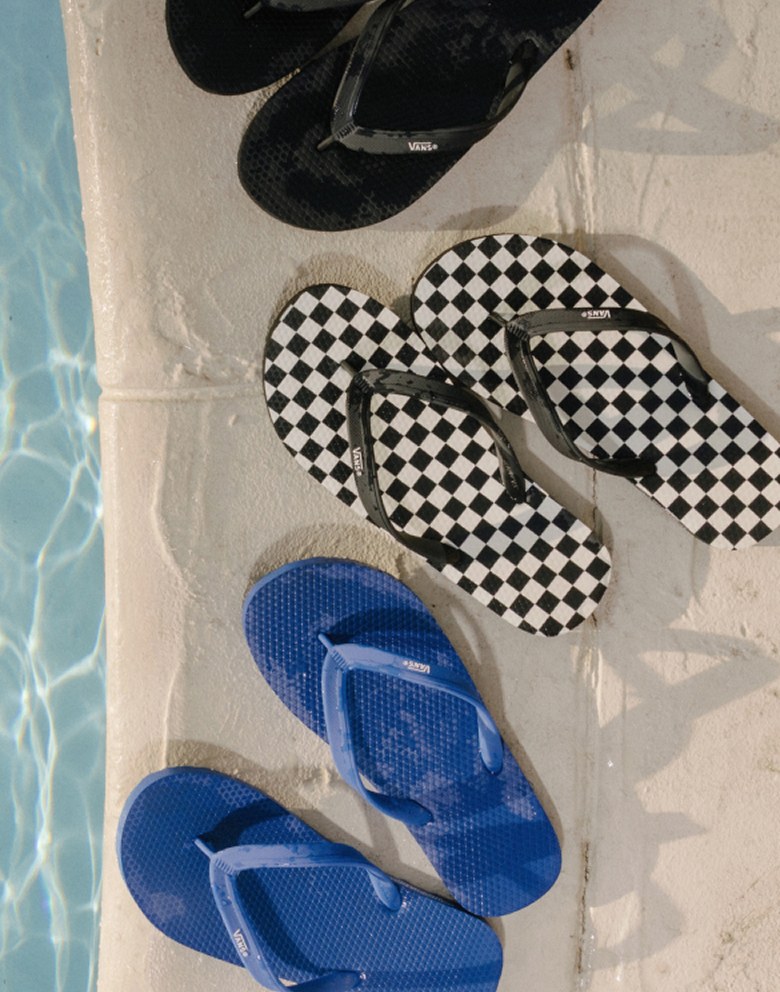 A black Soft-Top Sandal, a blue Soft-Top Sandal, and a black and white Makena Checkerboard Sandal sit on a pool deck next to the water.