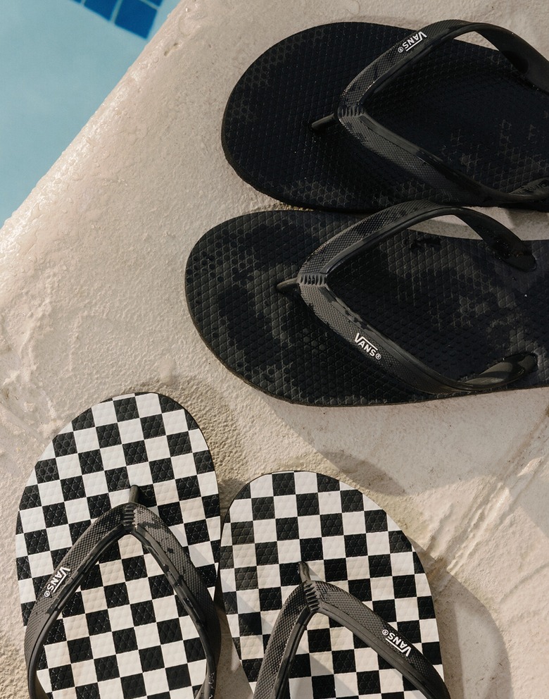 A black Soft-Top Sandal and a black and white Makena Checkerboard Sandal sit on a pool deck next to the water.