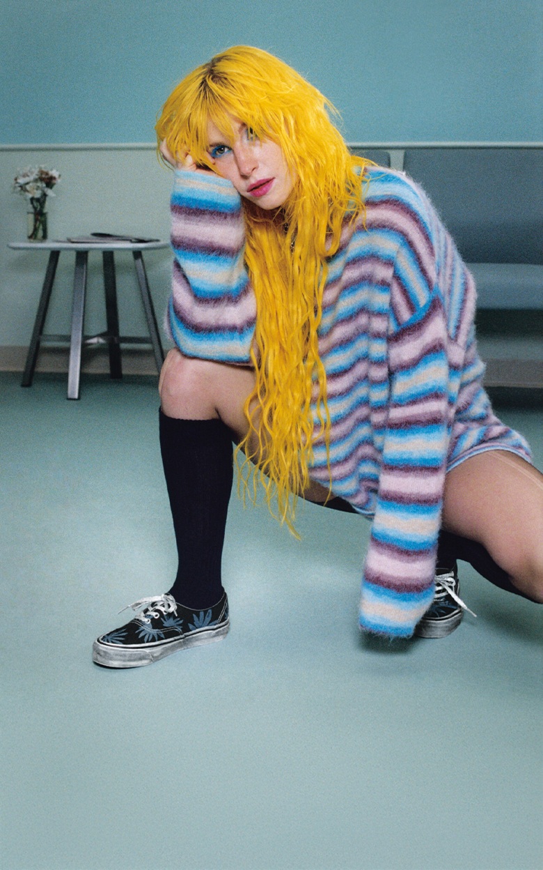 Hayley Williams, chanteuse du groupe Paramore, est accroupie dans une salle d’attente. Elle porte le Authentic à motif floral noir et bleu de la série Héritage Authentic pour la collection Porté haut et fort.