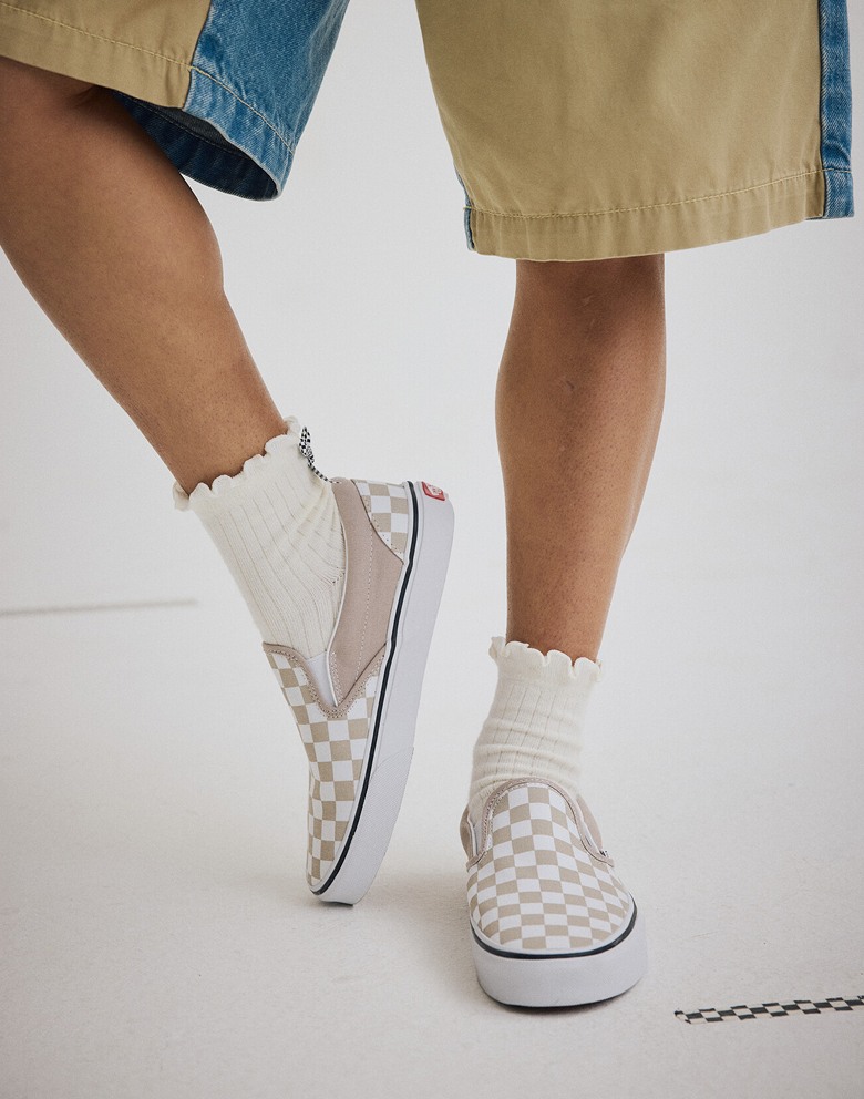 A knee-down shot of someone wearing mushroom brown Checkerboard Classic Slip-On Checkerboard shoes with white frilly socks in a photography studio.