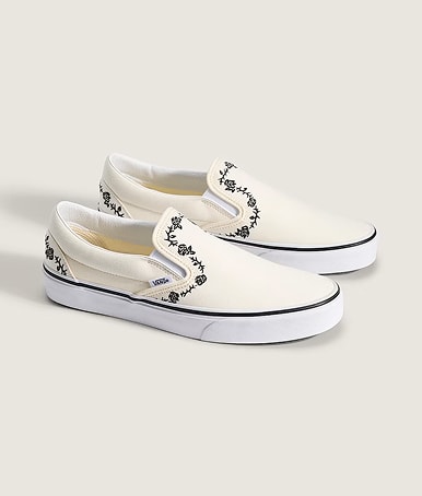 A pair of white Classic Slip-On with embroidered black floral details on the floor of a photography studio.
