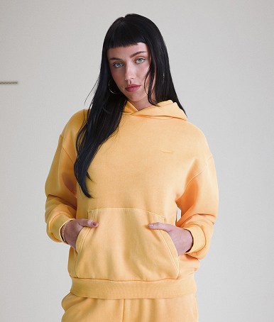 Une jeune femme porte un chandail à capuchon Salton et un pantalon de jogging droit en molleton Salton délavé couleur moutarde épicée dans un studio photo.