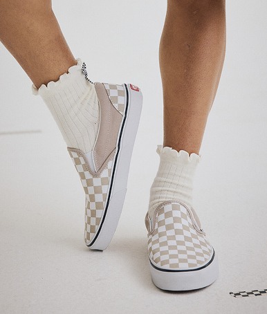 Plan à partir du genou d’une personne portant des Classic Slip-On à motif en damier brun champignon avec des chaussettes blanches à volants dans un studio photo.