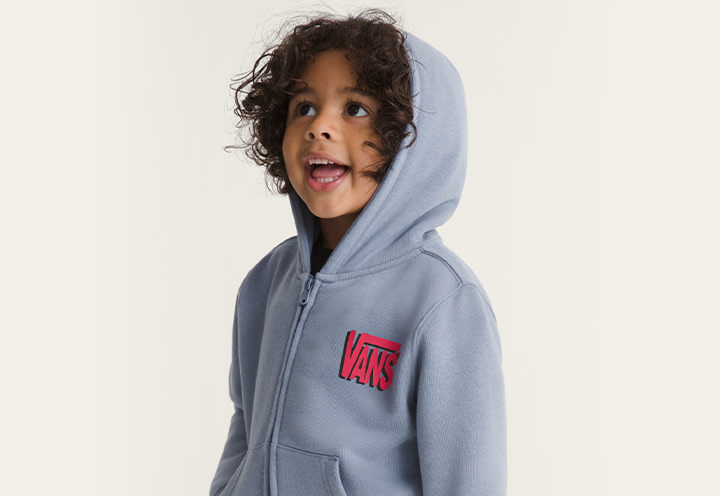 A young boy smiling and wearing a glacial slate blue Little Kids Yolo Penguin Zip Hoodie in a photography studio.