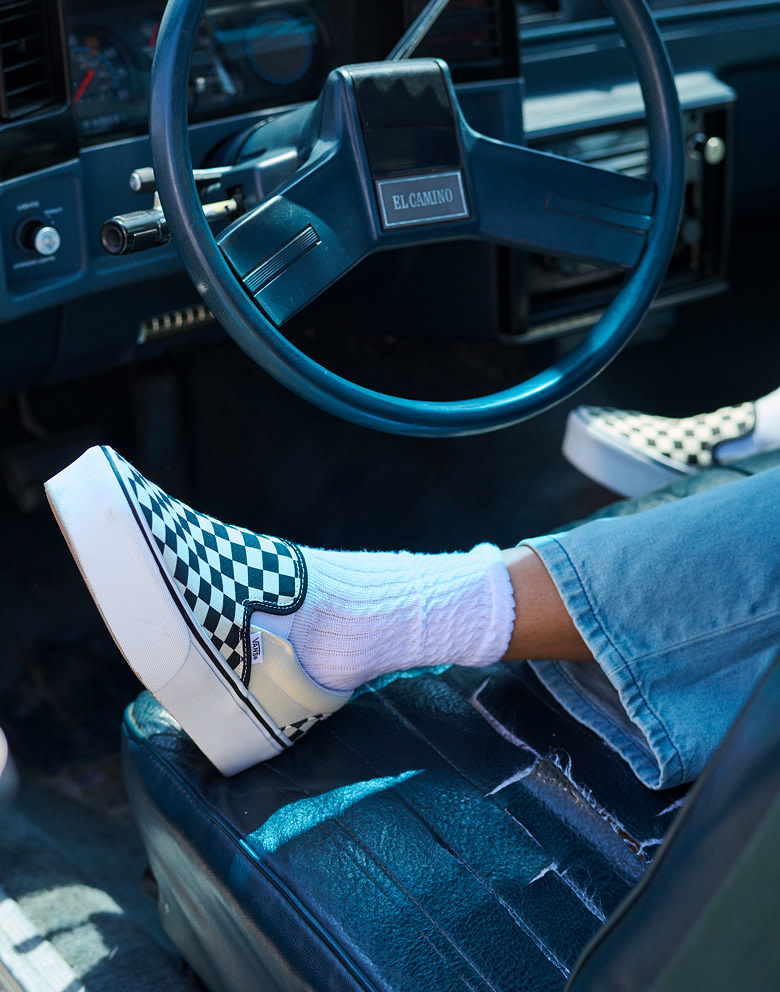 Close-up on a young woman wearing a black and white Classic Slip-On Platform Checkerboard shoe in a vintage car with Check-5 Low Rise Baggy Pants.