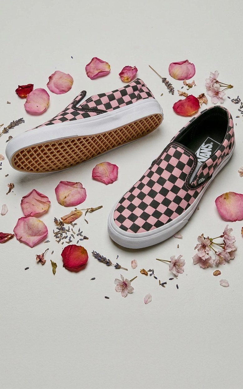 A pair of black and pink Checkerboard Classic Slip-On shoes on the floor of a photography studio surrounded by rose buds and petals.