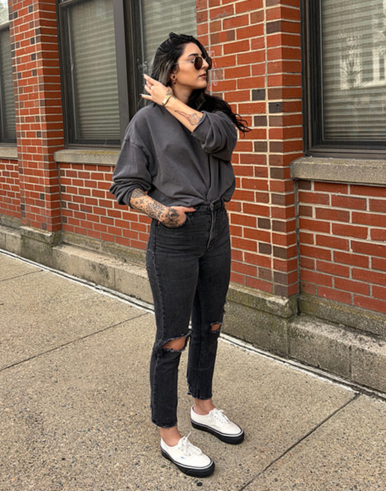 A young woman pulling back her hair on a sidewalk wearing white Authentic shoes, black jeans, and a black pullover tucked in.