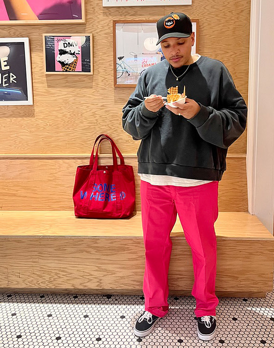 A young man eating ice cream against a wooden backdrop wearing black Authentic shoes, magenta pants, and a black crewneck.