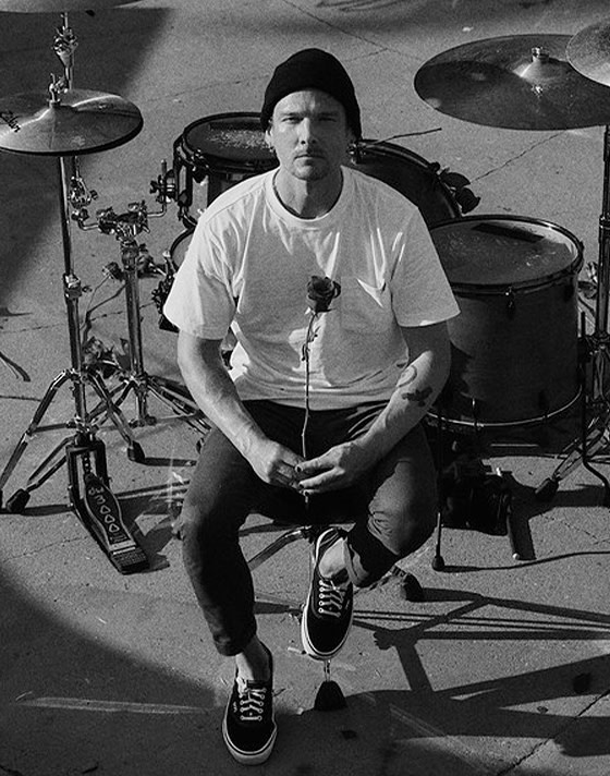 A black and white photo of a young man sitting next to his drum set and holding a rose in black Authentic shoes.