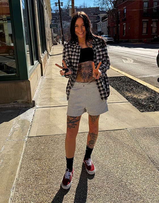 A young woman puts up peace signs wearing burgundy Authentic shoes, grey sweat shorts, and a flannel on a sidewalk.