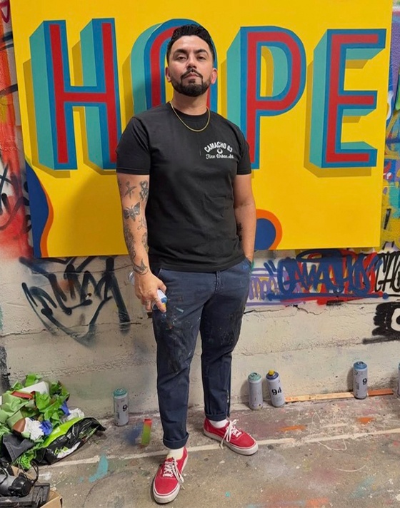 A young man holds a paint spray can and poses in front of his art wearing red Authentic shoes, paint-smudged blue pants, and a black t-shirt.