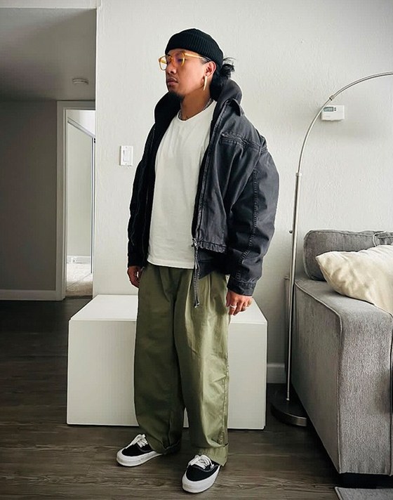 A young man stands in an apartment wearing black Authentic shoes, green baggy pants, a black jacket, and a black beanie.