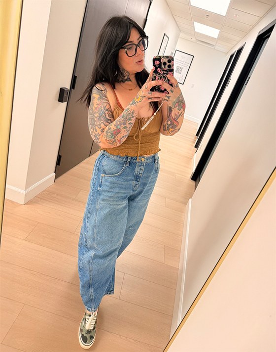 A young woman takes a selfie in a hallway wearing camo-printed Authentic shoes, blue jeans, and a dark gold tank top showing her arm tattoos.