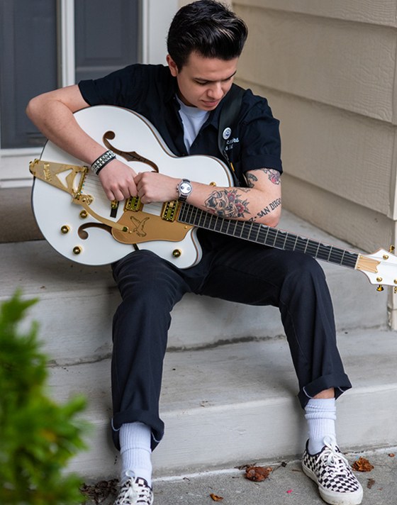 A young man sitting on a stoop and holding a white guitar wearing Checkerboard Authentic shoes with white crew socks and a full black fit.