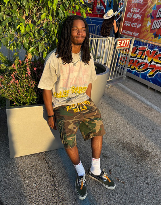 A young man sitting on a planter box wearing black Authentic shoes, camo shorts, and a white t-shirt at golden hour.