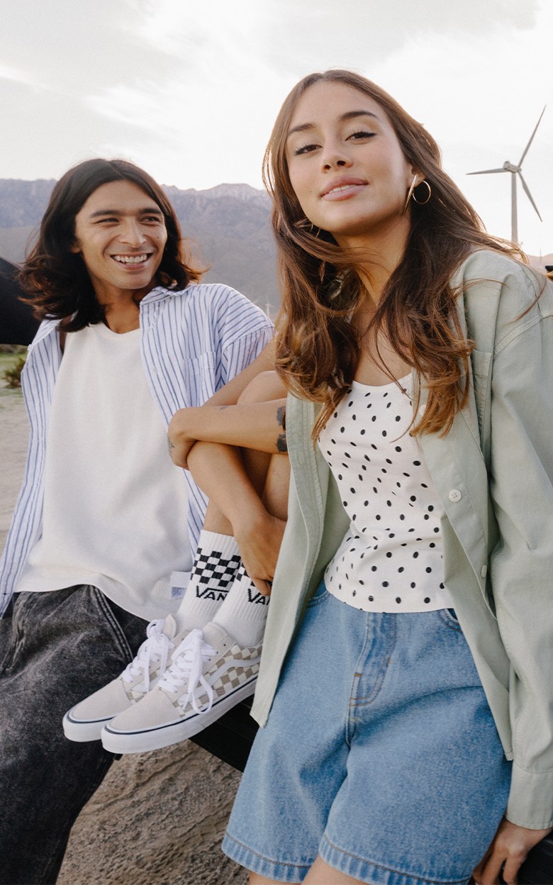 Medium panoramic shot of three people sitting and leaning on the tailgate of a truck in a windmill farm. All are wearing festival Vans gear.