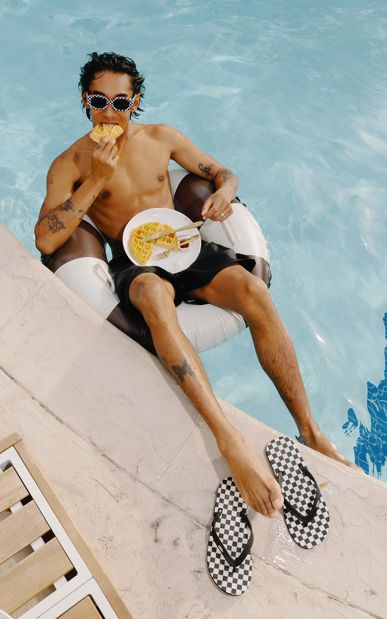 A young man floating in a pool eating waffles in Vans board shorts next to the Makena Checkerboard Sandal.