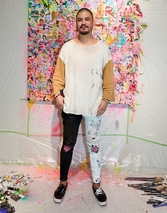 A young man poses in his art studio wearing black and white Authentic covered in paint.