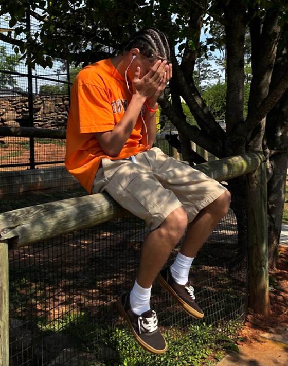 A young man sits on a wooden fence in the sun wearing black and gum Authentic shoes with brown shorts.
