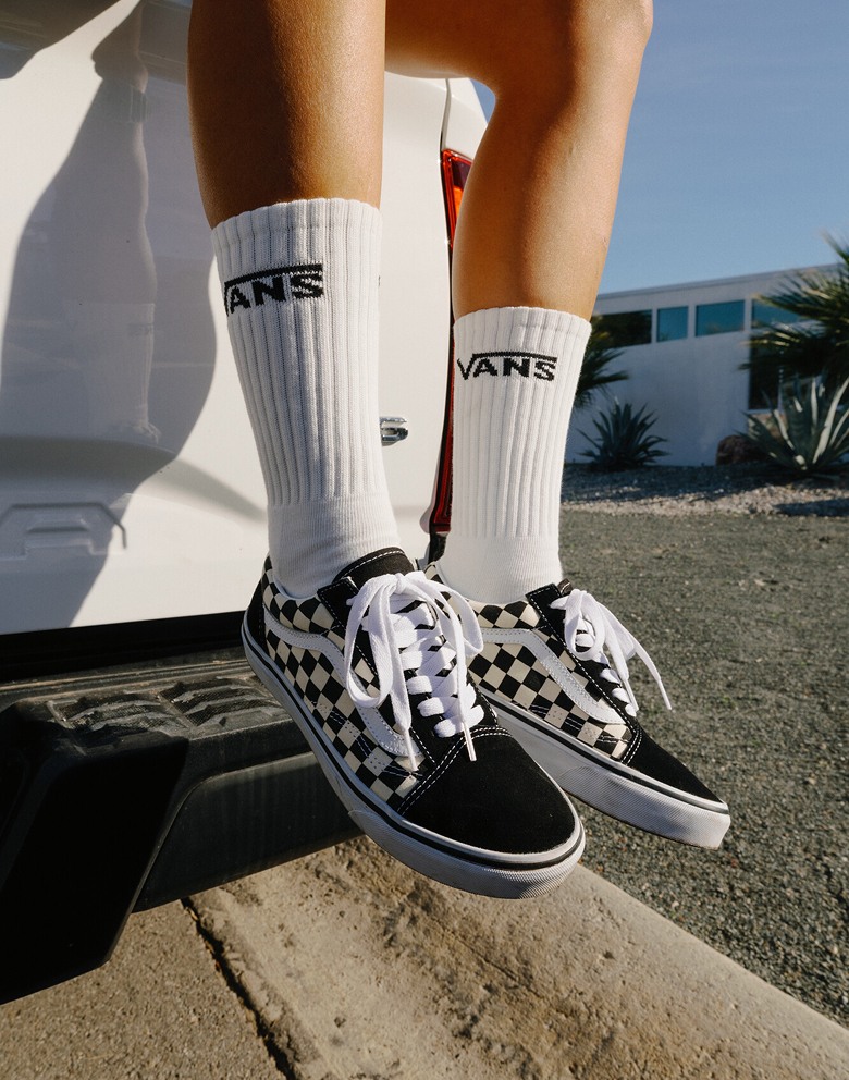 Close-up on someone sitting on a pickup truck and wearing black and white Checkerboard Old Skool with white Vans crew socks.
