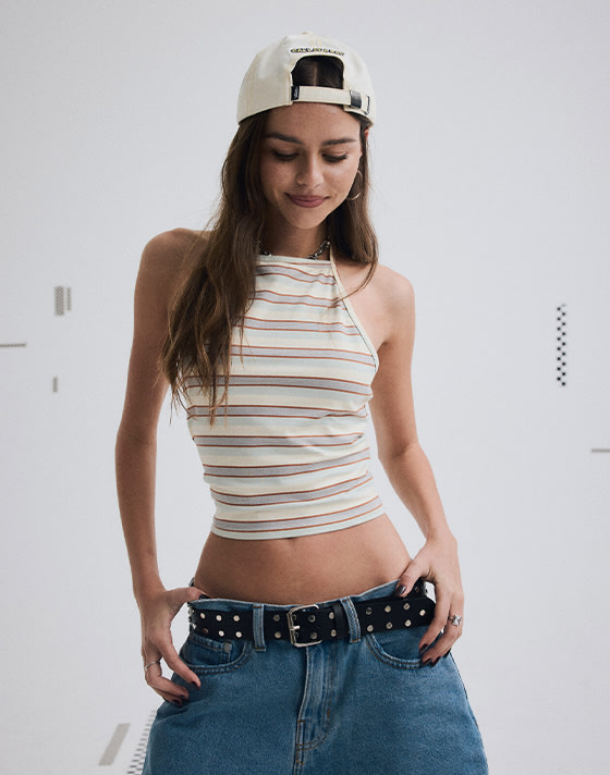 Medium shot of a woman from head to mid thigh. She's wearing a backwards hat with a striped crop top, baggy denim jeans and a thick black belt.