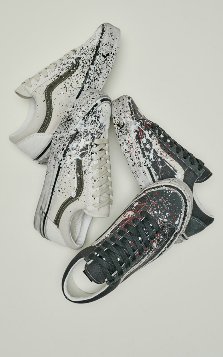 Two pairs of the paint splattered Old Skool on the floor of a grey photography studio. On the left, the white pair, on the right, a dark grey pair.