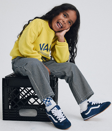 A young boy wearing black and white Super Lowpro shoes, a Little Kids Hot Dawg Crew Sweatshirt, and Little Kids Drill Chore Loose Stripe Pants.
