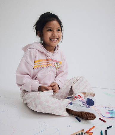 A young girl wears Little Kids Old Skool V shoes, pink Little Kids Lili Elastic Waist Checkerboard Pants, and a pink Little Kids Vans Sun Zip Hoodie.